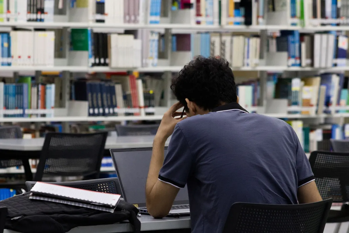 A person seen from behind sits at a desk with a laptop and notebook in a library, resting their head in their hand while surrounded by tall bookshelves
