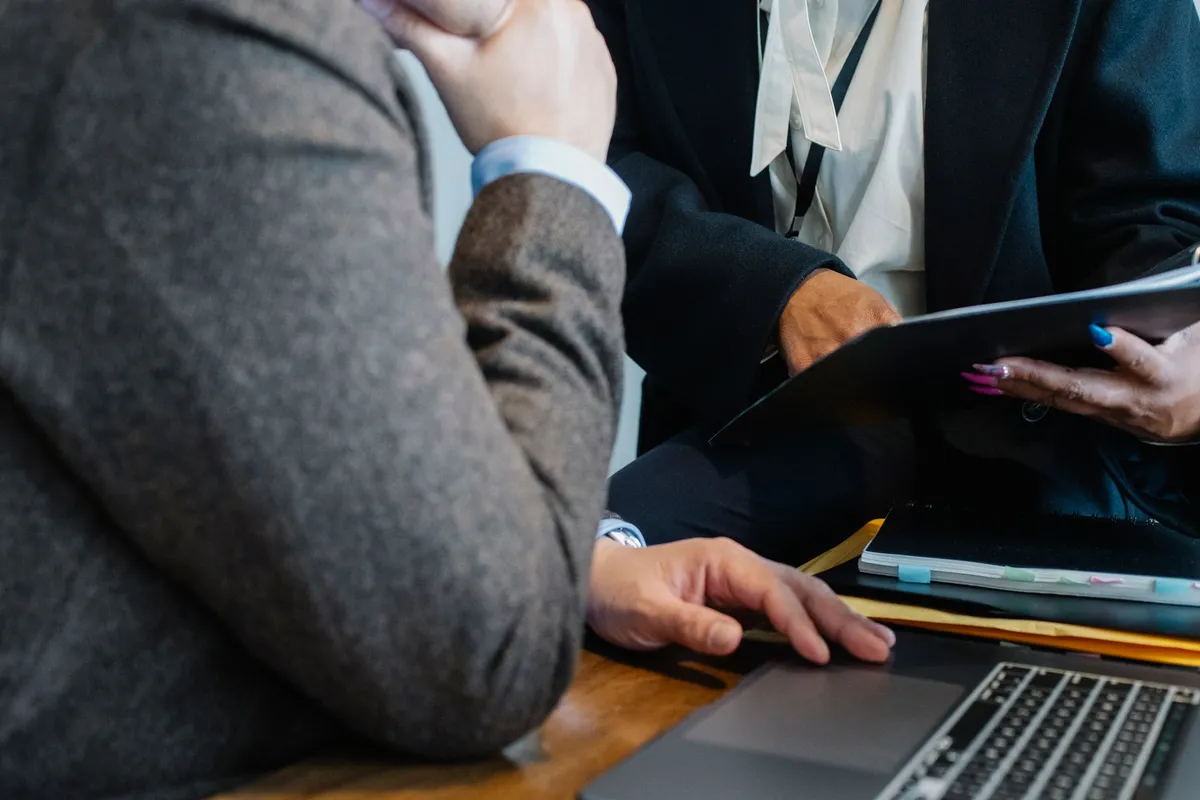 Close-up of people in business attire seated at a table with a laptop, notebook, and folder, one holding a tablet
