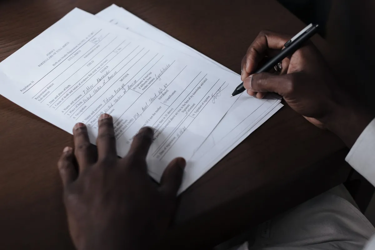 A person’s hands signing a birth and death registration form with a pen on a wooden desk