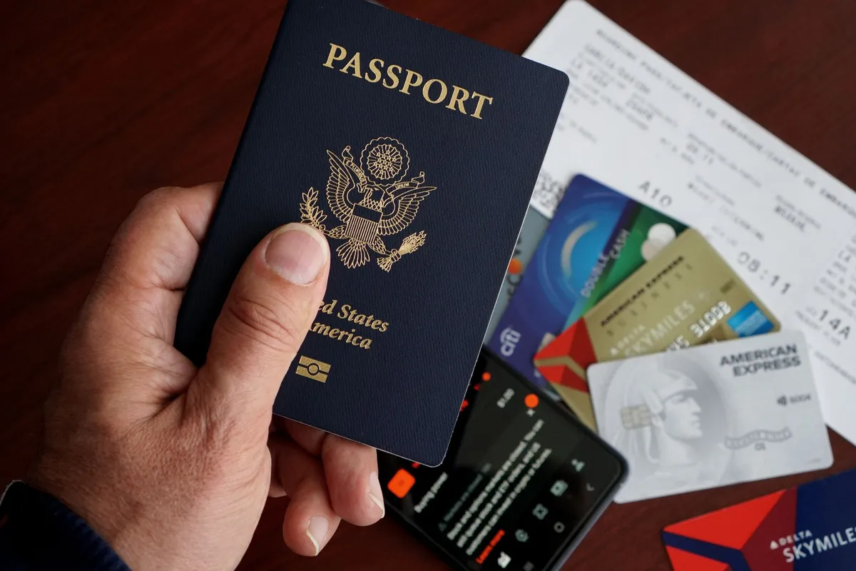 A hand holds a United States passport over a spread of credit cards, a smartphone, and a boarding pass on a wooden table