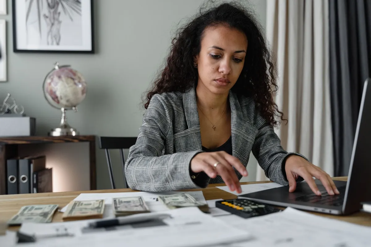 Woman in a blazer typing on a laptop at a desk with stacks of US dollar bills, a calculator, and paperwork in front of her