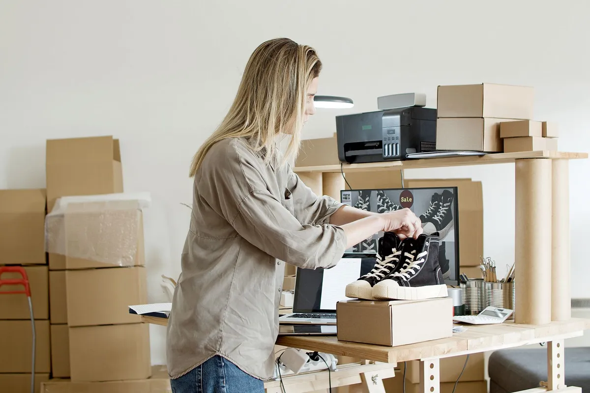A woman ties the laces of black and white high-top sneakers on a shipping box at a desk with a laptop, computer monitor showing a shoe sale image, printer, and stacks of cardboard boxes around her