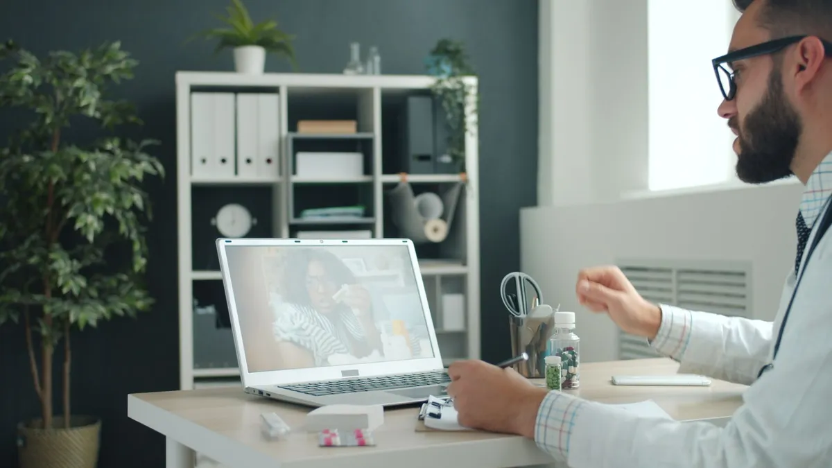 A bearded man in a white shirt with a stethoscope sits at a desk with medication bottles, gesturing while video calling a woman on a laptop screen