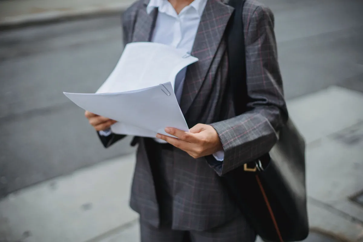 A person in a plaid blazer stands outdoors holding a paper-clipped stack of blank documents while carrying a shoulder bag