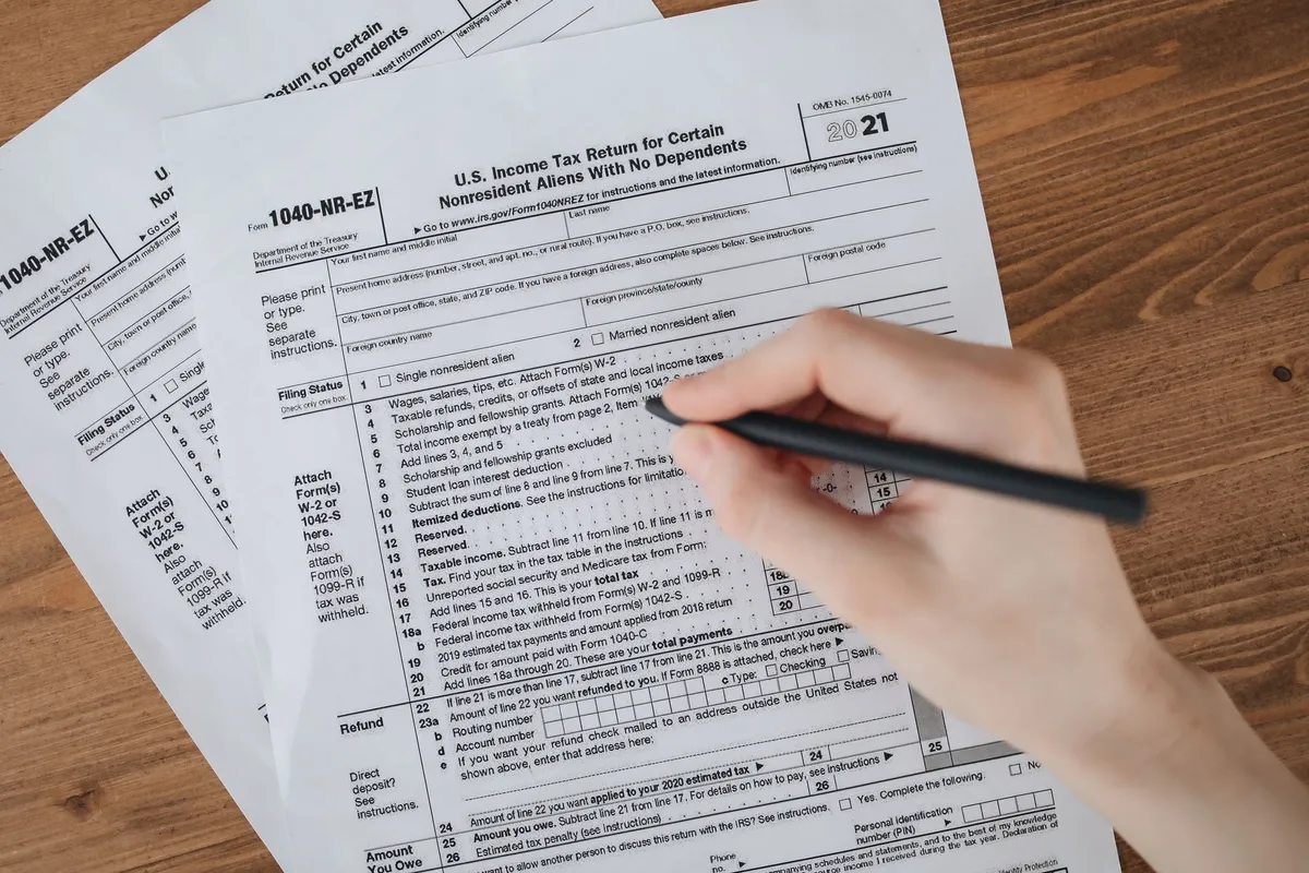 A hand holding a pen fills out a printed IRS Form 1040-NR-EZ U.S. Income Tax Return on a wooden table with additional copies of the form underneath