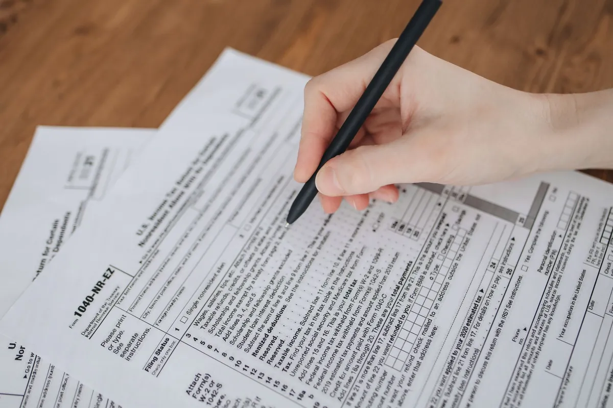 A hand holding a black pen fills out a U.S. Form 1040-NR-EZ tax document