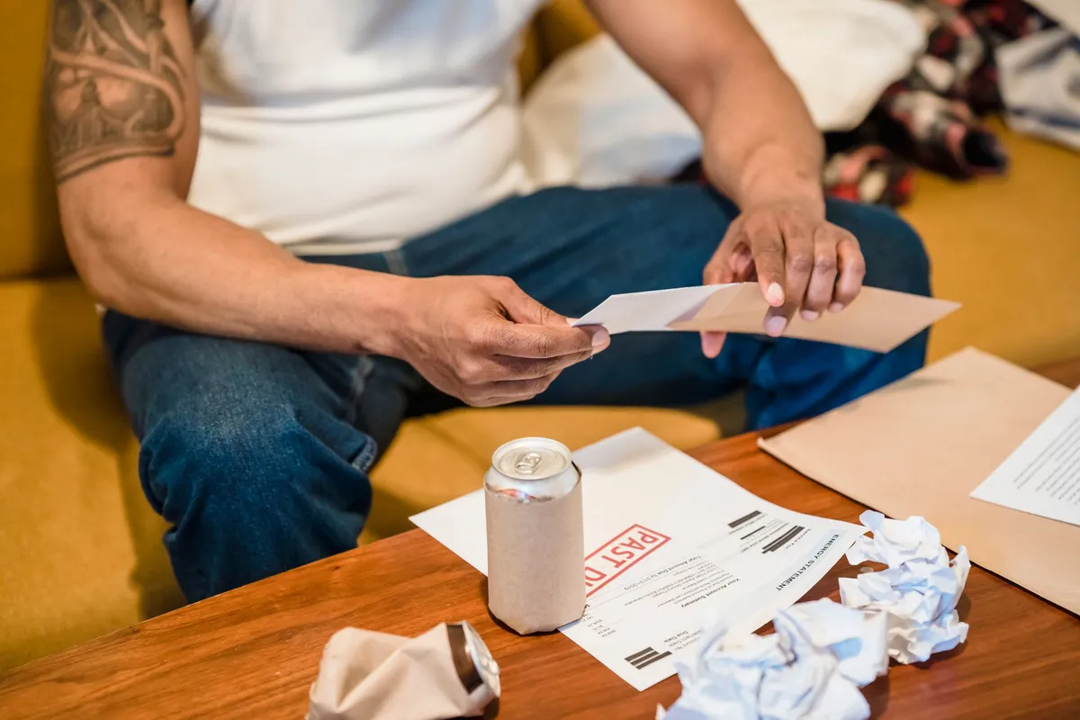 A tattooed person sitting on a couch opens an envelope near a coffee table with crumpled papers and a bill stamped PAST DUE