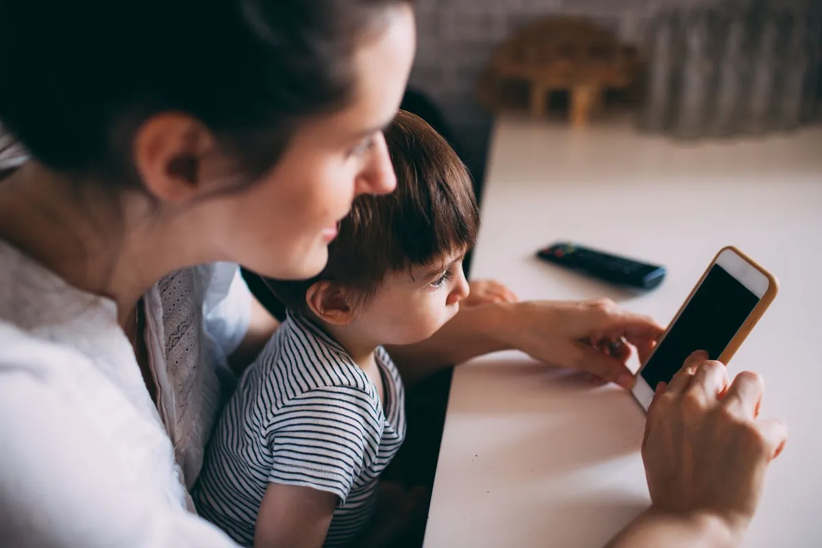 A woman holds a smartphone with a dark screen while a young child sitting on her lap reaches toward it, with a remote control visible on the table