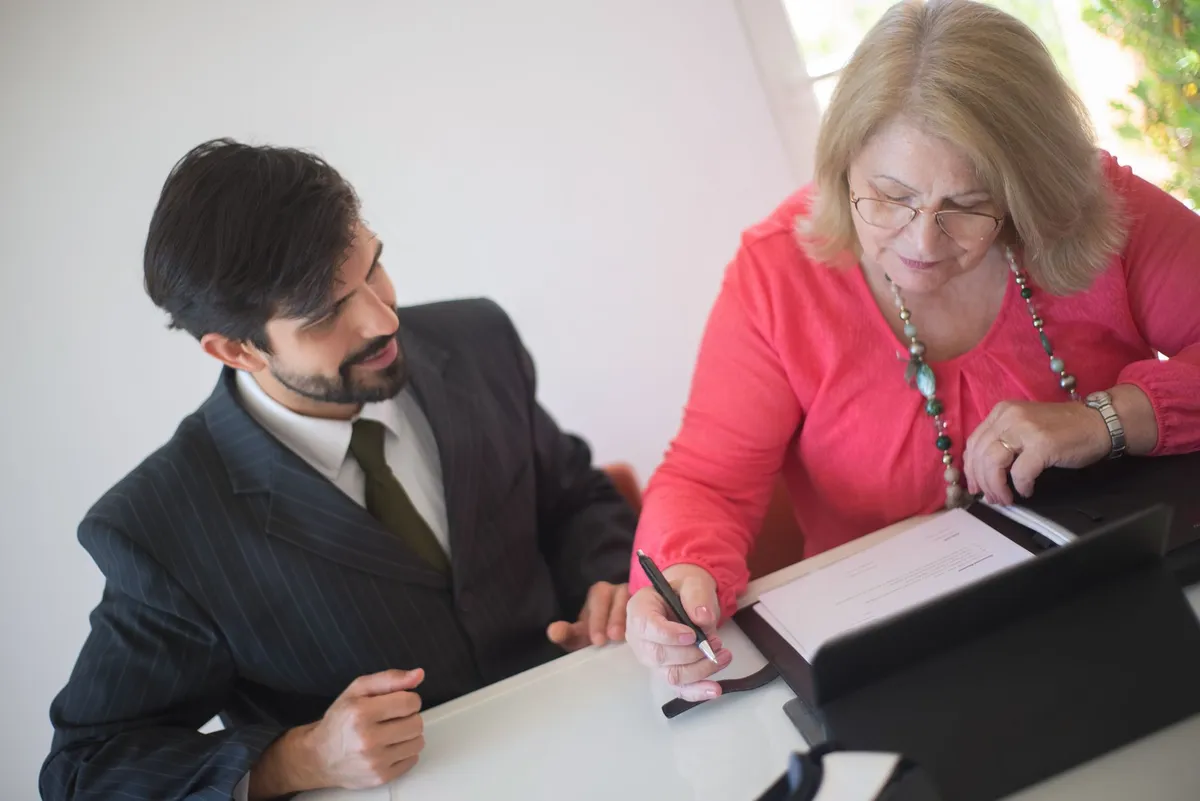 A man in a pinstripe suit sits beside a woman in a pink top who signs a document with a pen at a desk
