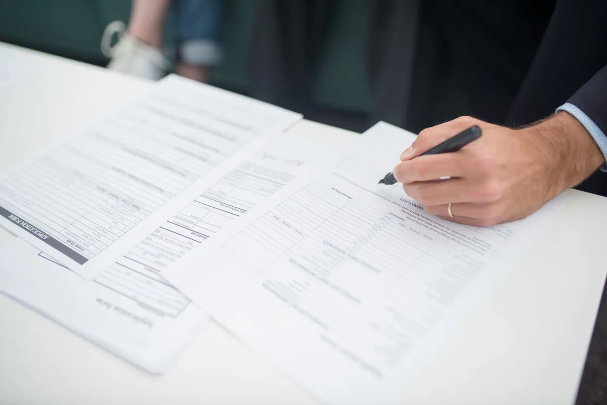 A person in a suit signs an application form with a pen, with additional paperwork spread on a white table