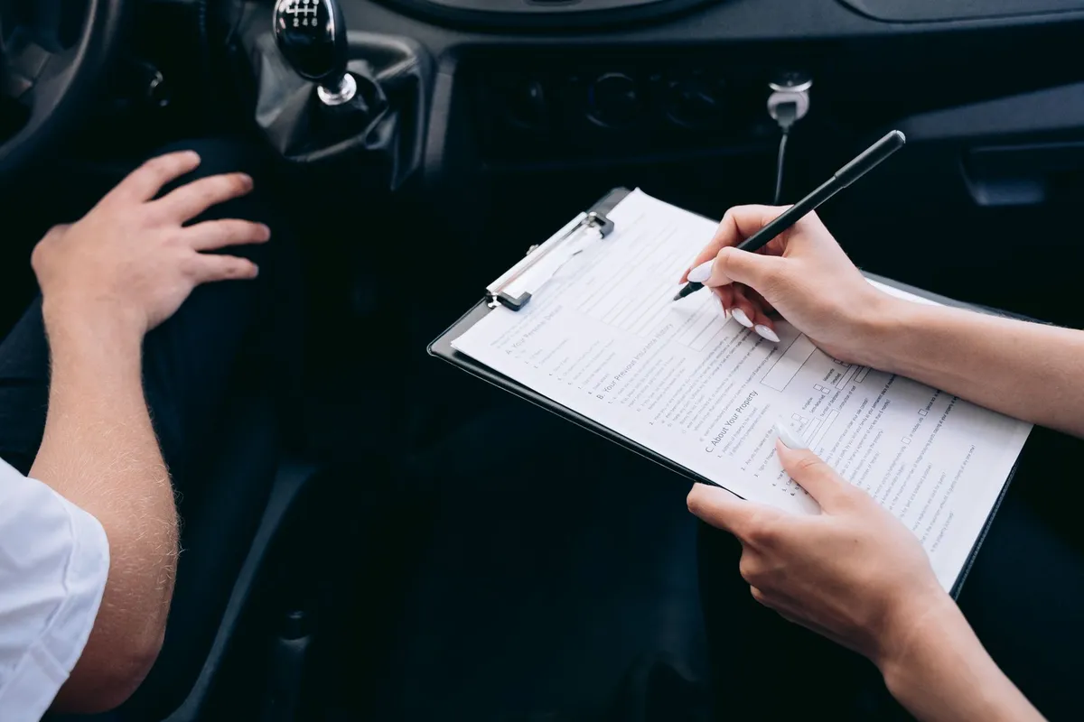 Inside a car, one person holds a clipboard with a printed form and writes on it with a pen while another person’s hand rests on the gearshift area