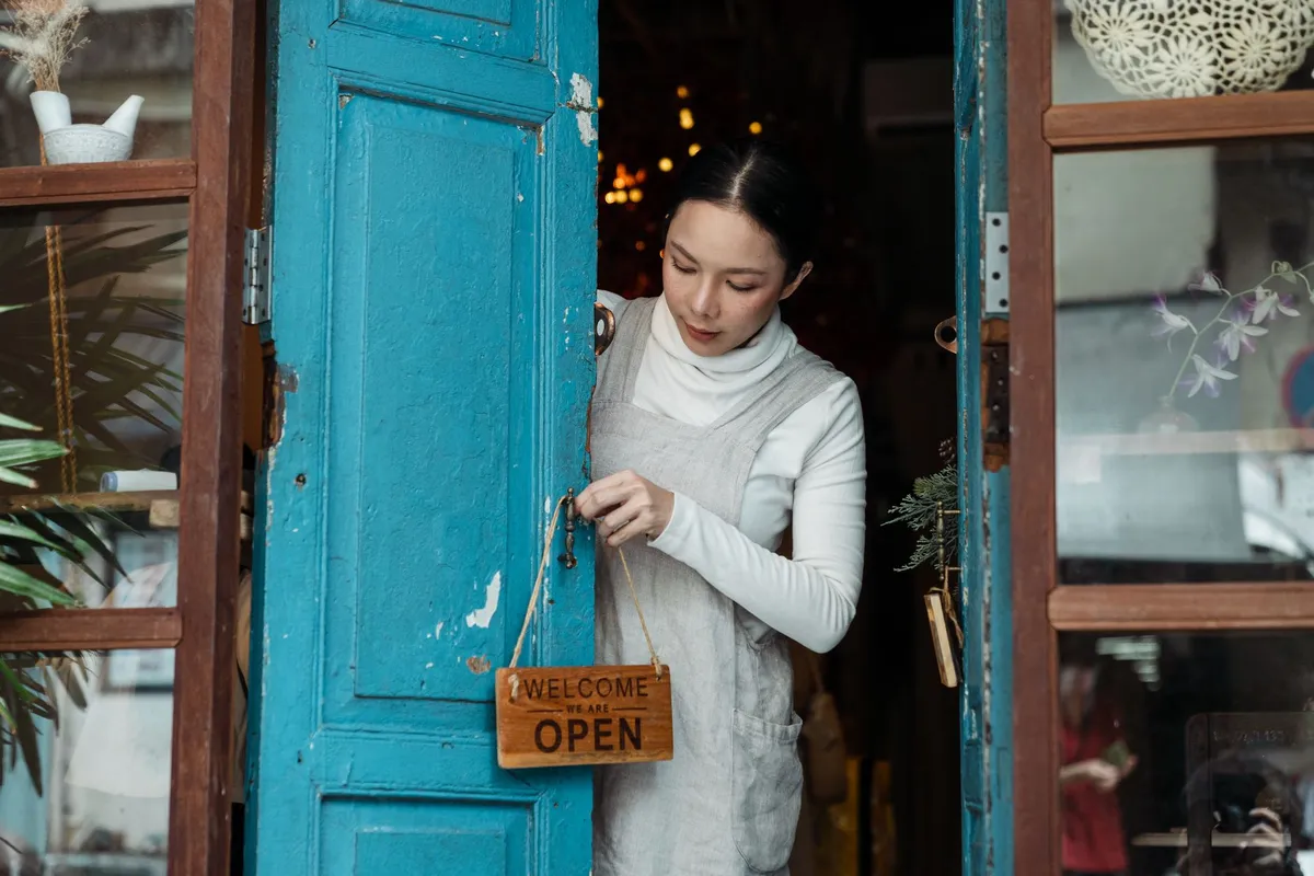 A woman hangs a wooden “Welcome We Are Open” sign on the handle of a weathered blue shop door
