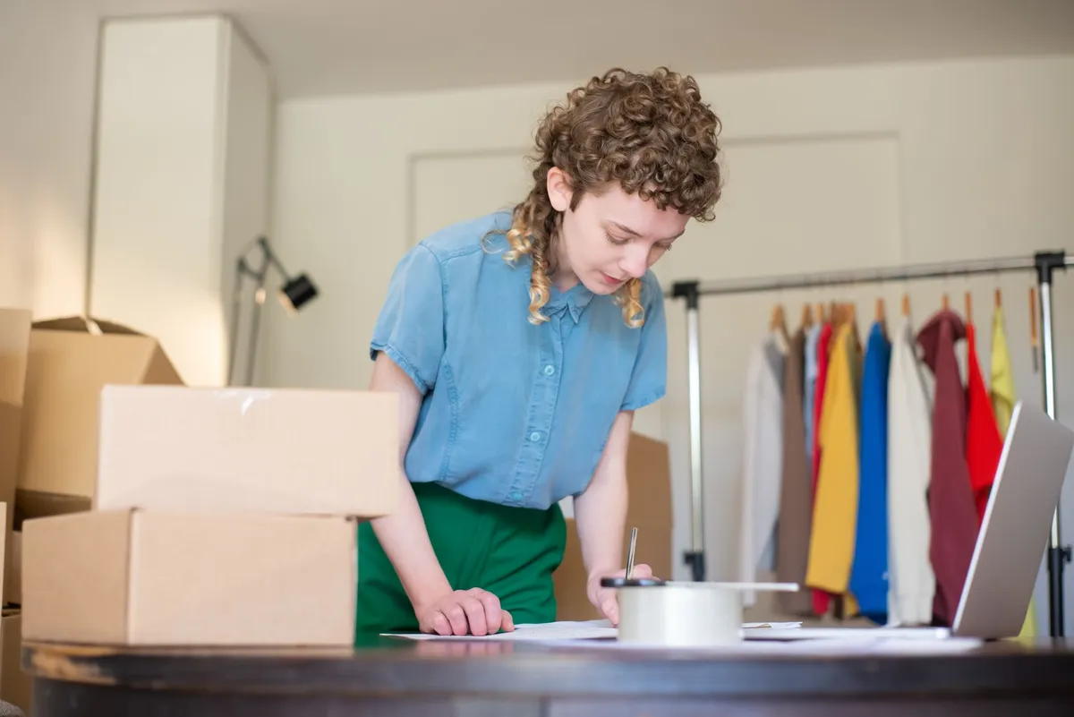 A woman with curly hair leans over a table stacked with cardboard boxes and a tape dispenser, with a rack of colorful clothing and a laptop nearby