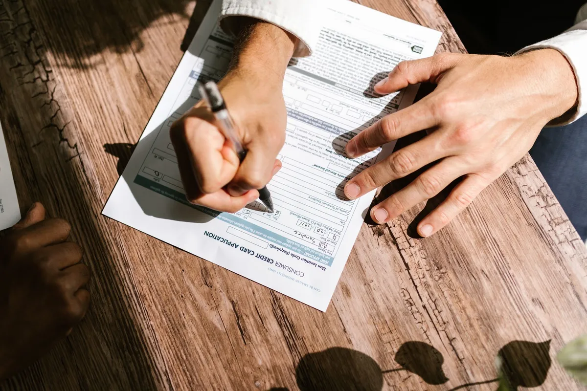 A person filling out a consumer credit card application form with a pen while another hand rests nearby on a wooden table