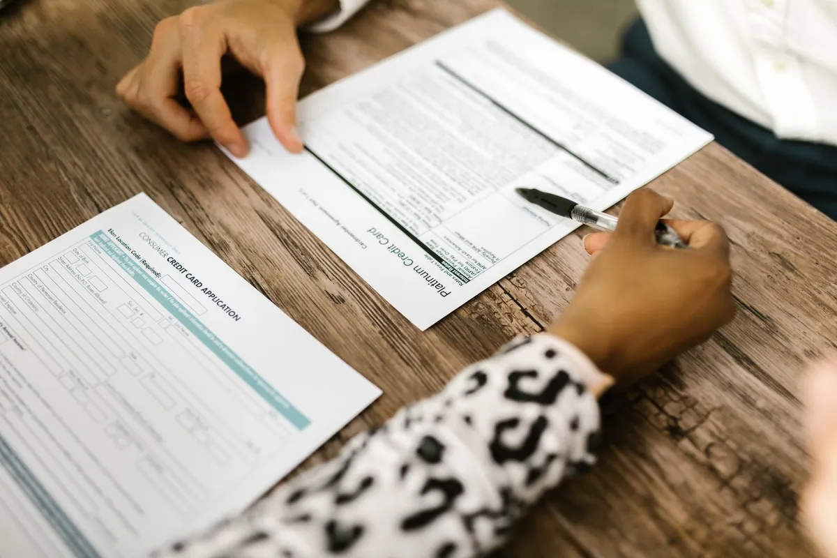 A person holds a pen over a consumer credit card application form while another person reviews a related document at a wooden table