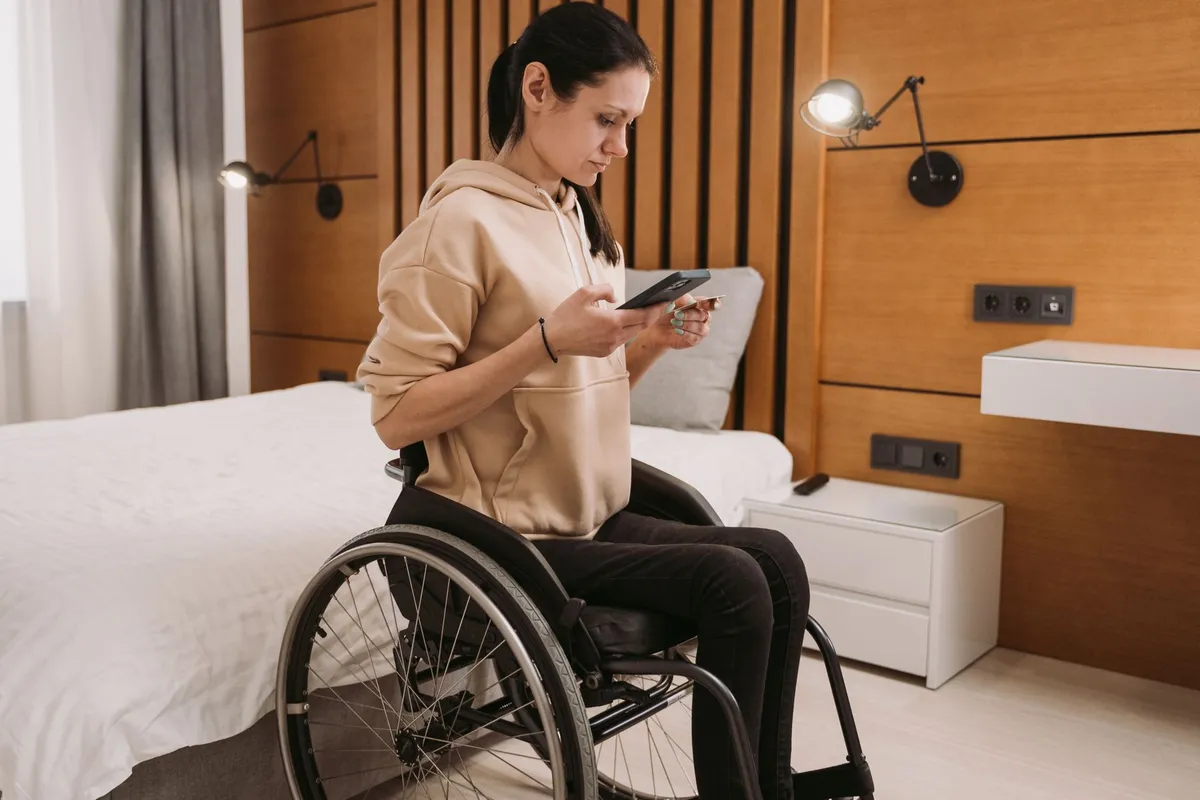 A woman in a wheelchair wearing a beige hoodie looks at her smartphone while sitting in a bedroom