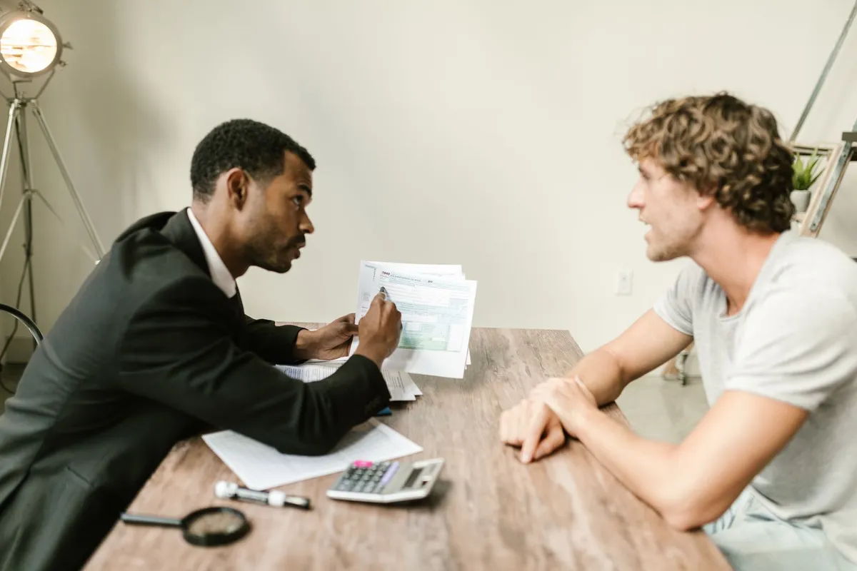 A man in a dark suit holds up a tax form while seated across a wooden table from a man in a white t-shirt, with a calculator and magnifying glass on the table between them