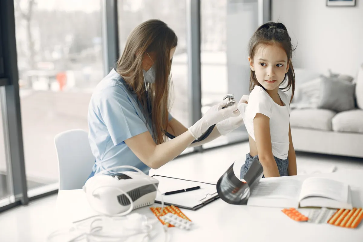 A masked healthcare worker in blue scrubs uses a stethoscope to examine a young girl’s back while she sits on an exam table surrounded by medical equipment and medication blister packs