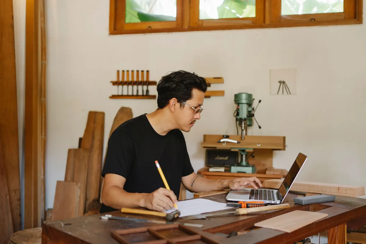 A man in a woodworking workshop writes on paper with a pencil while using a laptop at a cluttered wooden workbench surrounded by tools and a drill press
